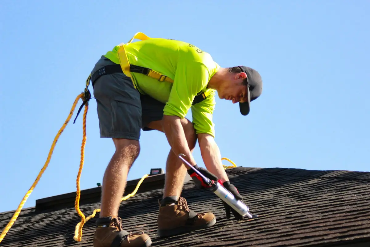 A Brannock roofer in a safety harness sealing flashing on a shingle roof under a clear sky