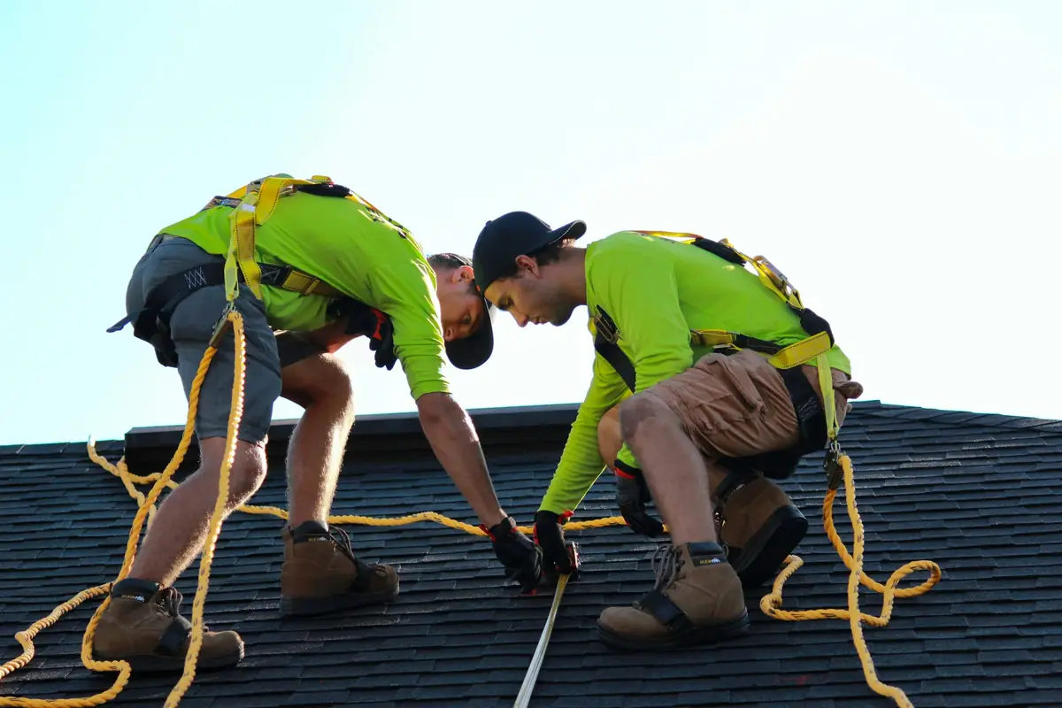 Two roofers kneeling on a roof, measuring and nailing shingles
